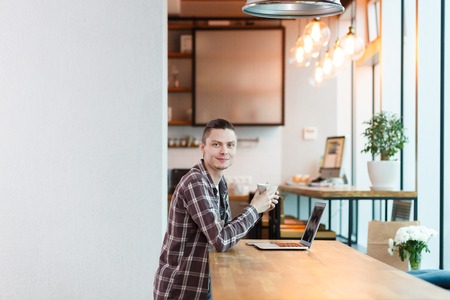 Side view - young handsome manager looks at camera and drinks coffee while working on sturtup project in an empty studio apartment in beautiful modern interior. Big city lifeの写真素材