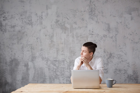 Cute young male writer romantically looking out the window while working on new scientific book sitting at the desk with laptop and books on the gray vintage background. Copyspaceの写真素材