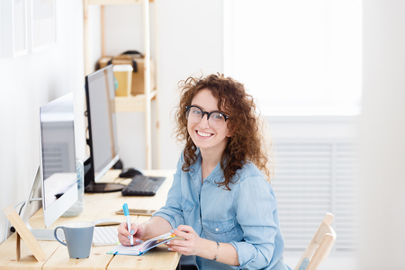 Indoor shot of young female startupper or cheerful caucasian freelancer work in light office, has good mood, ooks with pleasant smile in camera. People, emotions,businessの写真素材