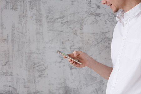 Side view of positive young man in casual clothes smiling and looking social network in smartphone on gray vintage wall background. Copyspaceの写真素材