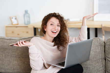 Attractive young woman chatts with friend on smart phone, surfing websites while shopping online at home, looking and smiling at the camera with broad smile.の写真素材