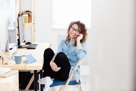 Charming glad caucasian female freelancer sitting at working desk at home and doing remote job. Successful marketing expert holds smart phone as communicates remotely with partnersの写真素材