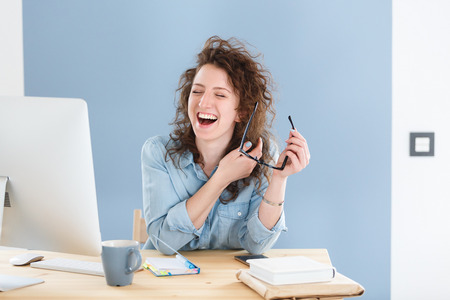 Funny employee of the company with curly red hair is pleased at his workplace in the office, has broad smile,sits opposite computer at the desk in creative studioの写真素材