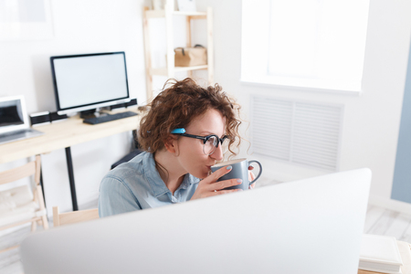 Young nice girl designer sits at a desk in office in front of computer and drinks hot coffee, takes a short break during work day, enjoys chatting with colleagues. Creative professionの写真素材