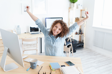 Young pleasant employee of the company happily finished the working day. Sits at the desk and smiles at the camera. Concept of success, work, peopleの写真素材