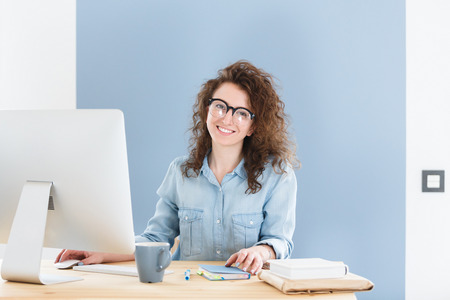 Cheerful European business woman in round spectacles, smiles happily sitting at office and studies business documents, looking and smiling at the camera during working day.の写真素材