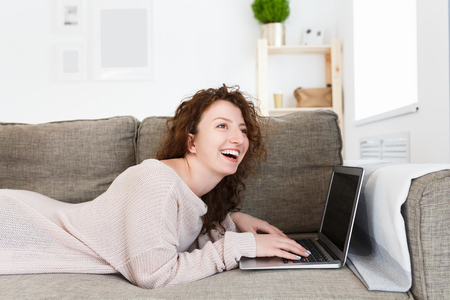 Happy positive young successful female lying on sofa, chatts with friend on laptop computer, surfes internet. Woman demonstrates positive emotions or feelingsの写真素材