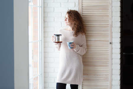 Beautiful caucasian woman with curly red hair drinking coffee,standing by the big window, enjoying a quiet cozy evening at homeの写真素材