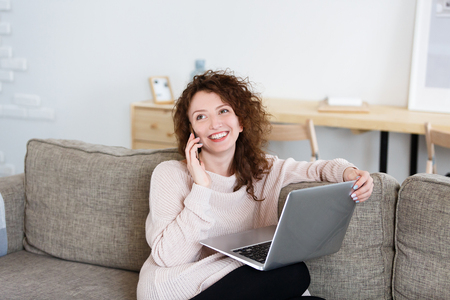 Pretty smiling glad female dressed casually sitting in front of open laptop in cozy living room and talking on phone with someoneの写真素材