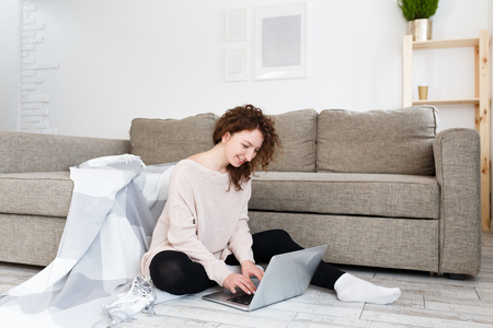 Young student girl writes a diploma on laptop. Ð¡harming female is surfing high-speed internet for articles useful for the project, sitting on the floor with a soft blanket. Education concept, lifestyle, technologyの写真素材