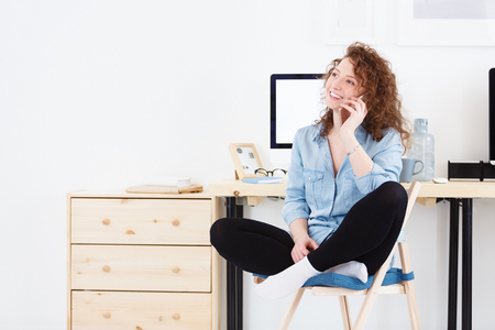 Beautiful young caucasian female with curly red hair in jean clothes talks happily with relative from sweet home. Remote work in another countryの写真素材
