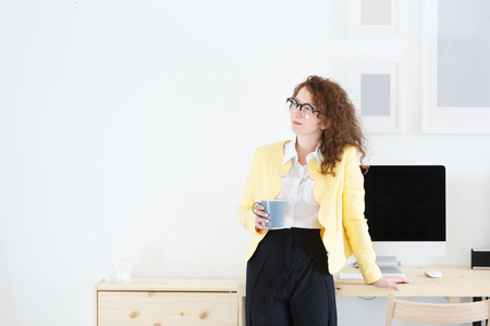 Horizontal portrait of gorgeous beautiful young successful female manager in white modern office, stands at the desk and drinks coffee during break of working day.の写真素材