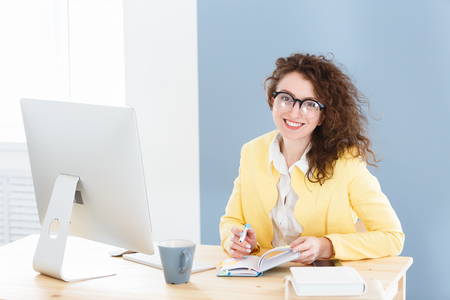 Attractive young female entrepreneur with red hair wearing stylish glasses and casual yellow jacked sitting at desk after hard working dayの写真素材