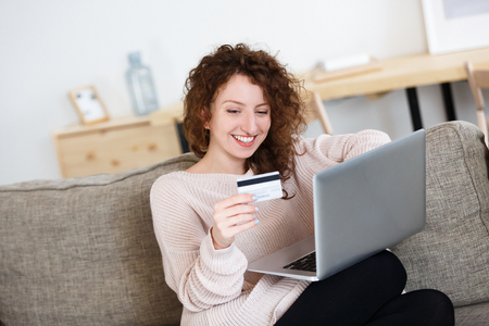 Good-looking woman holds credit card and modern laptop pc, surfing websites while shopping online, pays the restaurant delivery of food at home. Modern technologiesの写真素材