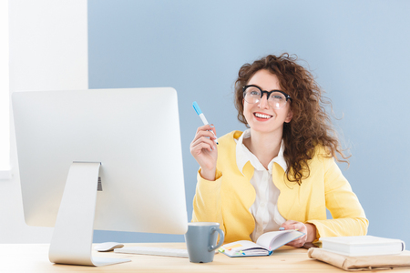 Happy positive young successful journalist has overjoyed expression, sitting at desk in front of computer and making notesの写真素材