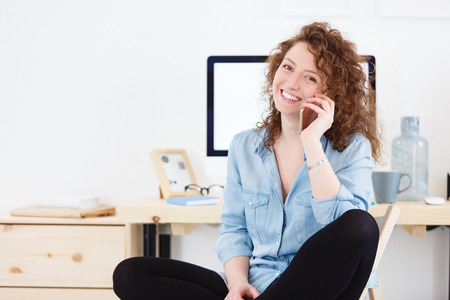 Pretty smiling glad female dressed casually sitting in front of desk with digital computer, talking on phone with friend after hard work dayの写真素材