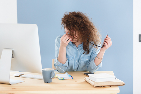 Sleepy businesswoman sitting at white office desk with electronic devices, holding eyeglasses, rubbing his eyes and massaging nose bridge, has fatigue expression over blue wall backgroundの写真素材