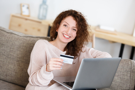 Happy and joyful redhead woman looking at camera with happy expression, hold in hand credit card, making transaction using laptop pc. Online shopping. People, business and finances concept.の写真素材