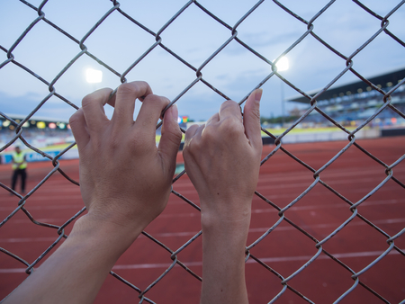 Close up on a woman's hands and  man's hands as They are holding on to a fence outside a Football field.の写真素材