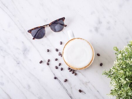 Top view of hot Cappuccino in the white cup ,coffee bean and black sunglasses put on the fabric and marble table in cafe morning work day background.の写真素材