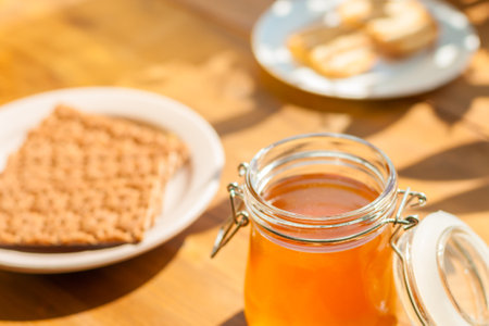 A glass jar filled with golden honey rests on a wooden table, while a plate of crackers and cookies compliments the inviting sunlight ambiance.の写真素材