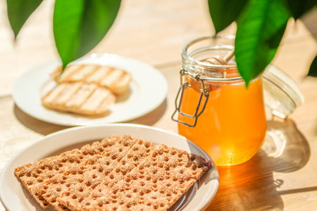 Matzah bread rests on a plate beside a jar of honey in a sunlit kitchen, showcasing a calm and inviting atmosphere with light filtering through green leaves.の写真素材