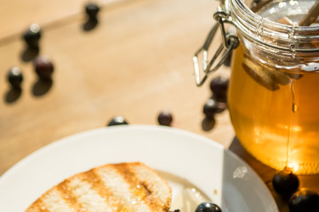 Grilled toast garnished with honey and sprinkled with fresh blueberries sits on a rustic wooden table, illuminated by warm sunlight, evoking a cozy breakfast vibe.の写真素材