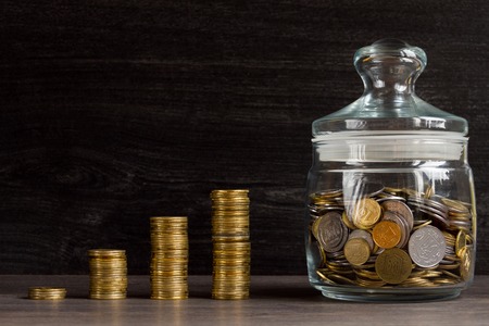 Save money concept. Stack of golden ukrainian coins and glassy moneybox on wooden background