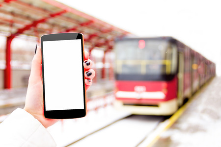 Female / woman / girl hand with mockup smartphone / mobile phone with blue tram and public transport station on background in winterの写真素材