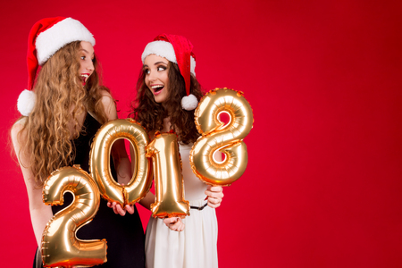 Joyful and happy young females / women in Santa hat holding gold balloons numbers 2018 as symbol of...