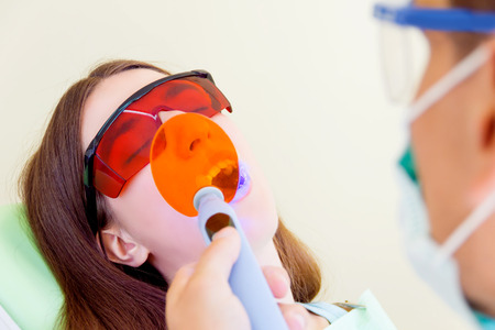 Young man male dentist in medical glasses and mask treatments teeth of woman female patient in red uv glasses by uv equipment tool implanting medical sealantsの写真素材