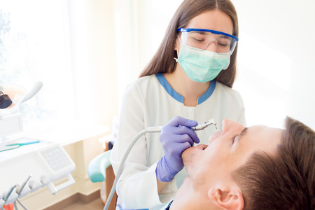 Young woman female dentist in medical glasses and mask with dentist equipment tool machine preparing for treatments teeth of smiling man male patientの写真素材