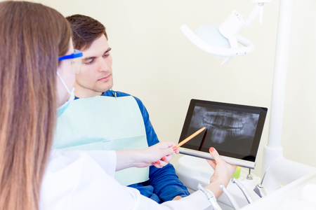 Young professional dentist woman female in white grown, medical mask and glasses with x-ray photo of patient teeth on tablet explains treatment for male man in dentist chairの写真素材