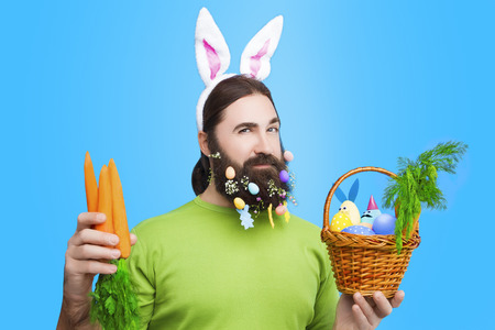 Happy Easter concept. Nice kind muscle man male with beard, white ears of rabbit, carrots and basket with colorful easter eggs and green in green t-shirt isolated on blue backgroundの写真素材