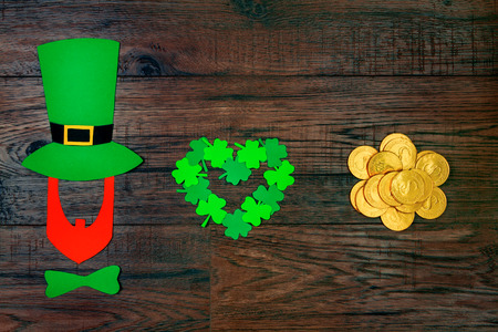 Saint Patrick's Day. Silhouette of leprechaun in green hat and green tie bow with green three petal clover in shape of heart and gold coins on wooden backgroundの写真素材