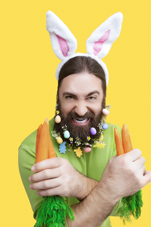 Happy Easter concept. Nice kind muscle man male with spring's flower's beard, white ears of rabbit, carrots in green t-shirt isolated on yellow backgroundの写真素材
