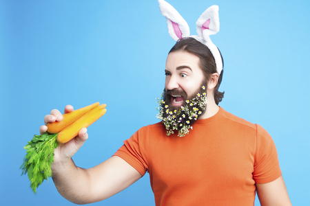 Happy Easter concept. Nice kind muscle man male with spring's flower's beard, white ears of rabbit, carrots in orange t-shirt isolated on blue backgroundの写真素材