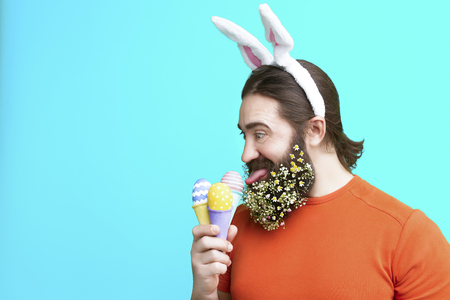 Happy Easter concept. Nice kind muscle man male with flower's beard  with white ears of rabbit and colorful easter eggs in waffle in orange t-shirt isolated on blue backgroundの写真素材