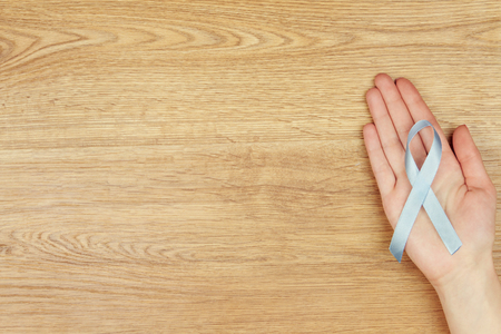 Oncological disease concept. Woman's hand with a light blue ribbon as a symbol of prostate cancer isolated on wooden background.の写真素材