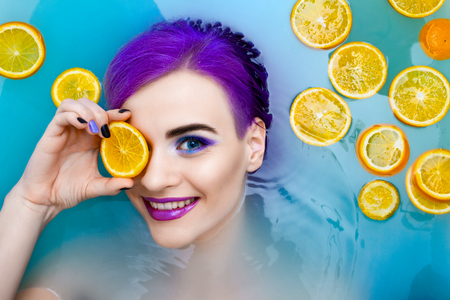 Portrait of young pretty female girl adult woman / fashion luxury model with purple hair relaxing in bathtub with oranges isolated by blue water. Organic skin care, beauty and body care concept.の写真素材