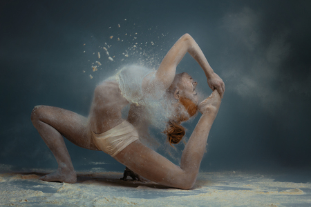 Dancing in flour concept. Redhead beauty gymnast female girl adult woman dancer in dust / fog. Girl wearing white top and shorts making dance element in flour cloud on isolated grey black backgroundの写真素材