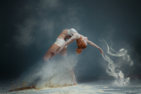 Dancing in flour concept. Redhead beauty female girl adult woman dancer in dust / fog. Girl wearing white top and shorts making dance element in flour cloud on isolated grey black backgroundの写真素材