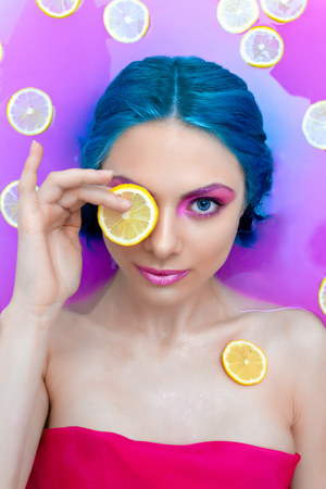 Portrait of young pretty female girl adult woman fashion luxury model with blue hair and bright make up relaxing in bath with lemons oranges isolated by pink water. Organic skin care, beauty and bodyの写真素材