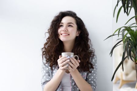 Portrait of young pretty girl woman female wearing pajamas sleepwear with white hearts sitting near white wall drinking cup of coffee tea and smiling in the morning. Morning wake up conceptの写真素材