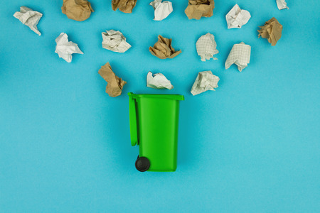 Paper and brown craft paper garbage trash and green trash bin with wheels on blue isolated background. Environment issue / care, recycling / reuse, save planet, ecology concept.の写真素材
