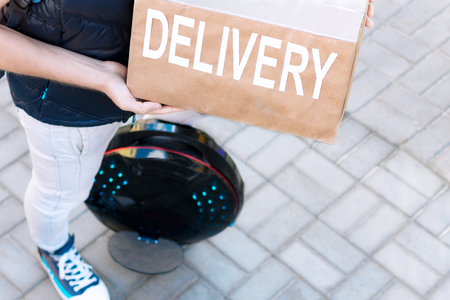 Modern future delivery. Man male guy in delivery uniform and delivery box with electric transport / eco ecology electric unicycle / self balancing wheel delivers ordered package to receiverの写真素材