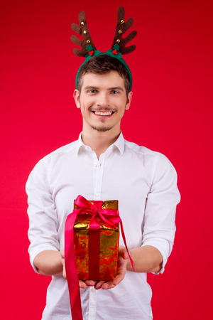 New Year party concept happy fun smiling charming handsome hipster man guy male celebrating winter Christmas holidays wearing white shirts 
deer horn hat holding give presents box gift red backgroundの写真素材