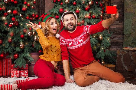 Happy family couple in love with santa and elf hats warm sweaters doing selfie with smartphone for greeting parents with Merry Christmas and Happy 2019 New Year at home with decorated christmas treeの写真素材