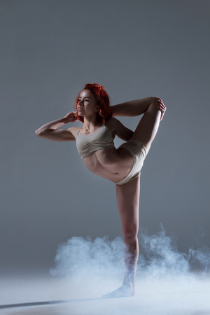 Dancing in cloud concept. Muscle redhead beauty female girl adult woman dancer athlete gymnast in dust fog smoke wearing dance clothing making dance element performance on isolated grey backgroundの写真素材