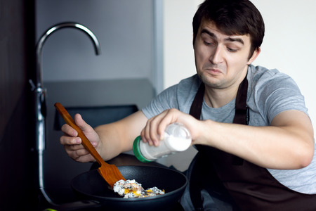 Young loser man male cooking scramble adding salt on metal frying pan in modern loft kitchen. Trying to make breakfast, lunch or dinner. Alone bachelor in kitchen without woman female conceptの写真素材